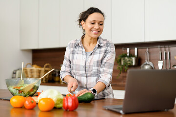 Cheerful young woman watching video blog about cooking. Female prepare salad, slices a vegetables...