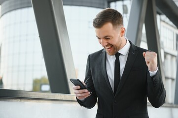 Fototapeta premium Portrait of an excited young businessman dressed in suit holding mobile phone, celebrating.