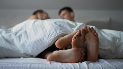 Close-up feet of unrecognizable blurred gay couple cuddling in bed in the morning. Happy carefree relaxed Caucasian young men enjoying leisure in bedroom indoors