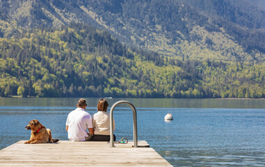 Couple with dog sitting on dock at the lake. Rear view of a couple sitting on pier with dog...