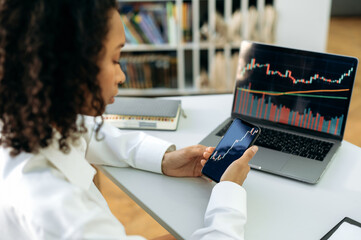 Confident smart dark-skinned girl, trader, stock investor, sits in office, uses smart phone and a laptop, works with quotes charts, analyzes the dynamics of the cost of crypto currency