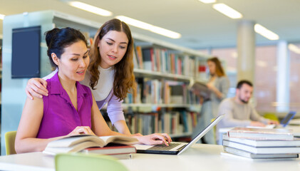 Two positive young women learning at library together, working with laptop and books