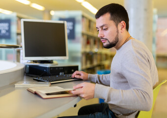 Young focused man reading book and working with computer, finding information and preparing to exam..at library