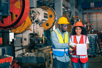 Two professional engineer/worker/technician use clipboard discuss work, walk in steel metal manufacture factory plant industry. Black African American man and woman wear hard hat check quality machine