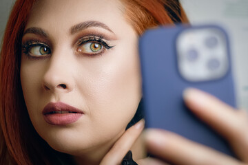 Blogger girl with a smartphone in her hand. Red-haired woman with bright makeup takes a selfie on the camera of a modern phone. Fashionable female hobby.