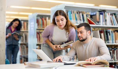 Male and female university students working on laptop computer in library, reading books and making...