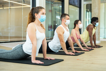 Group of sporty people in protective masks exercising pilates class in modern fitness center. Focus on young brunette. Healthy lifestyle and pandemic precautions