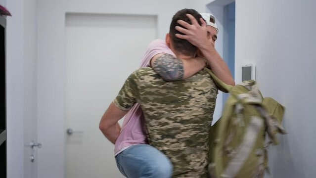 Happy gay soldier returning home as boyfriend jumping on hips hugging lover smiling. Excited Caucasian LGBTQ couple meeting indoors at home. Happiness and excitement concept