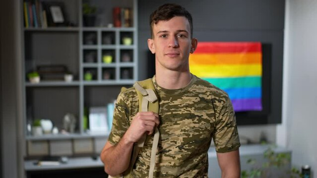 Portrait of confident young man in camouflage T-shirt looking at camera with rainbow LGBTQ flag at background. Brave Caucasian gay soldier posing indoors at home