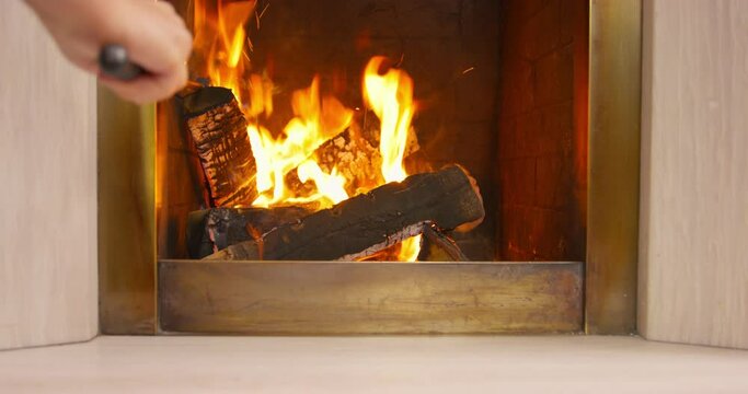A person straightens a wood log in the fireplace with a iron poker, to improve the flame. Cozy relaxing fireplace. Colorful flame, log burning on fire