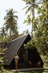 Sender woman in a white swimsuit stands in front of a bungalow in a tropical garden