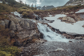 Mountain stream during spring flood, water breaks against rocks. Rivulet in mountainous area, rock ridge on horizon. Glaciers and snow on hillsides. Clouds over riverbed.