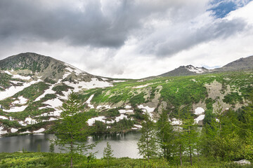 Stone ridge with glaciers and green shrubbery. Coniferous forest on shore of mountain lake. Journey through cliffs and cobblestones in spring, dangerous slope for descent on foot