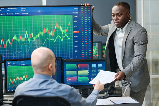 Unrecognizable mature man giving papers with financial stats of stock trading to his younger Black colleague
