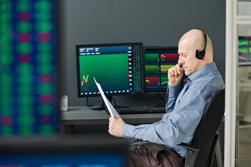 Mature professional Caucasian stock data analyst sitting at desk in office working with financial...