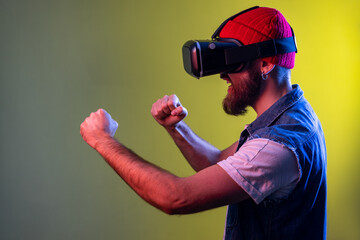 Side view of hipster man in virtual reality glasses on head playing fighting game, holding clenched fists up ready to boxing. Indoor studio shot isolated on colorful neon light background.