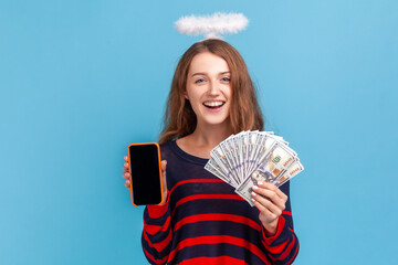Positive young adult woman wearing striped casual style sweater and nimb over head, showing smart phone with blank screen and dollar banknotes. Indoor studio shot isolated on blue background.