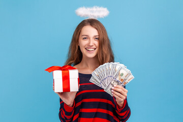 Optimistic woman wearing striped casual style sweater and nimb over head, holding present box and dollar banknotes, looking smiling at camera. Indoor studio shot isolated on blue background.