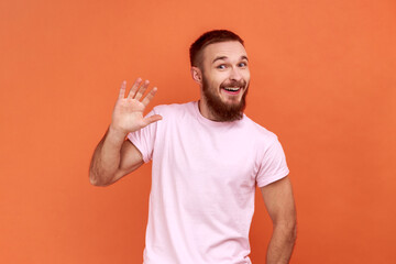Portrait of positive handsome bearded man waving hand and friendly smiling at camera, making hi...