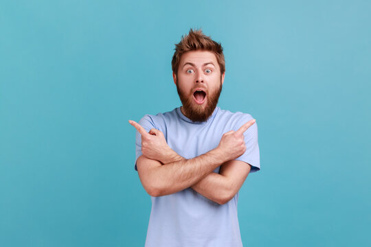Portrait of astonished surprised bearded man standing with crossed hands and pointing to copy space on both sides, presenting area for promotion. . Indoor studio shot isolated on blue background.