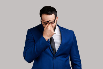 Bearded businessman standing with head down, rubbing eyes, putting off glasses, being tired work long hours, wearing official style suit. Indoor studio shot isolated on gray background.