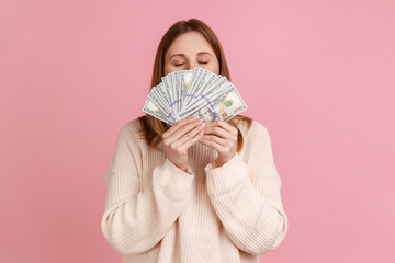 Portrait of rich greedy blond woman holding big fan of dollars bills and smelling money with satisfied expression, wearing white sweater. Indoor studio shot isolated on pink background.