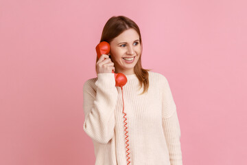 Portrait of happy satisfied blond woman talking on red retro telephone, having pleasant conversation, looking away with smile, wearing white sweater. Indoor studio shot isolated on pink background.