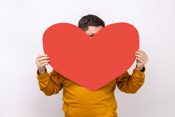 Portrait of man hiding face behind big red heart and looking at camera with curious prying eyes,...