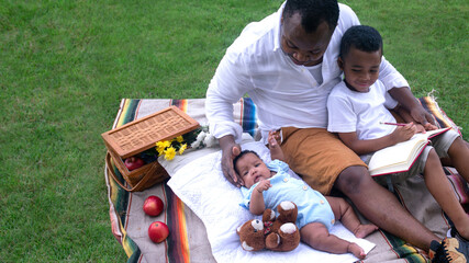 African father with lovely daughter and son having a great time, picnic on green grass in public...