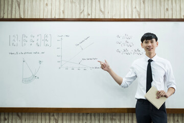 An Asian male teacher in a classroom near the whiteboard is writing and teaching a lesson with a...