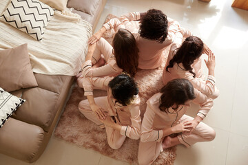 Young women in satin pajamas sitting in circle back to back and touching elbows when meditating or...