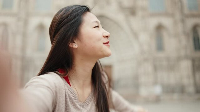 Brunette Asian girl sincerely smiling while taking selfie against the backdrop of old architecture. Japanese woman exploring new city