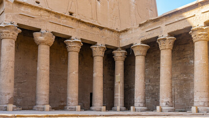 Naklejka premium Colonnade in the Temple of Horus in Edfu. Tall columns with various carved capitals stand in a row and in the corner of the hall. Hieroglyphs, drawings of gods and pharaohs are visible. Egypt
