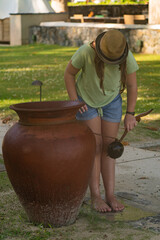 A girl wearing summer clothes washes her feet and legs on the beach using water from a big ceramic pot. 