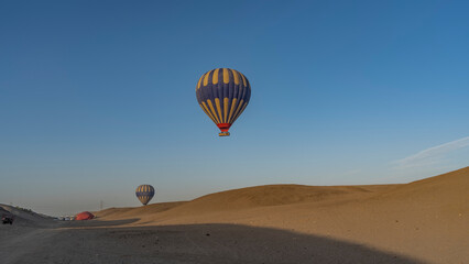 Bright balloons are flying in a clear blue sky. Below - the sand dunes of the desert. Several cars are waiting in the distance. Light and shadows. Egypt. Luxor