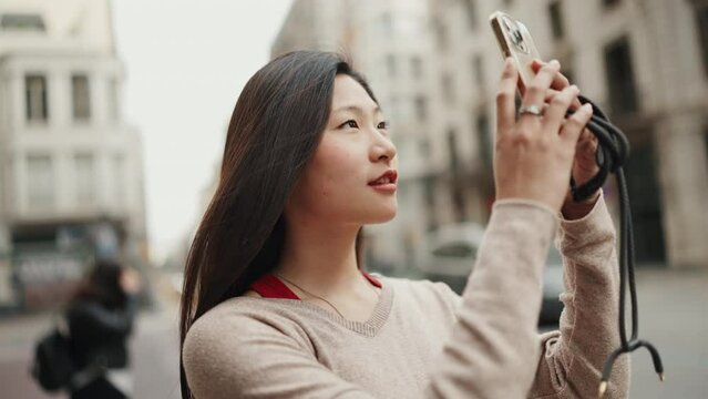 Attractive long haired Asian woman looking inspired taking photos of beautiful architecture on smartphone and smiling. Female Asian tourist exploring European city