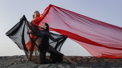 Two girls in black and red flowing fabric pose on sand dune