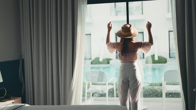 Rear back view woman opening white curtains enjoy and looking pool view, Happy traveller stay in high quality hotel.