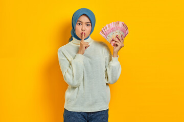 Beautiful Asian Muslim woman in white sweater, holding cash money in Indonesian rupiah banknotes, finger covering lips, makes silence gesture on yellow background. ​People religious lifestyle concept