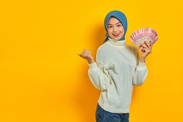 Cheerful beautiful Asian Muslim woman in white sweater  pointing thumbs at blank space, holding cash money in Indonesian rupiah banknotes over yellow background. ​People religious lifestyle concept