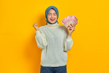 Excited beautiful Asian Muslim woman in white sweater holding cash money in Indonesian rupiah...