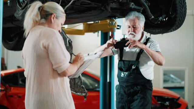Mechanic man making photo a damaged car after a collision, assesses and records the damage in a mechanic's workshop together with a female insurance agent