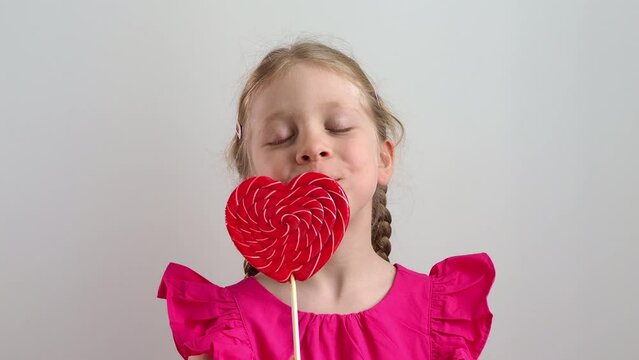 little girl with heart. The child holds a caramel heart in his hand, hides his face behind the heart. The girl congratulates on Valentine's Day and February 14. Baby eyes peek out from behind the cand