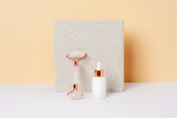 White serum bottle and crystal rose quartz facial roller, natural skin care products and tools on the natural stone background against yellow wall on white table