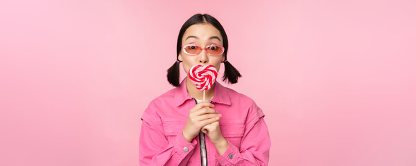 Stylish korean girl licking lolipop, eating candy and smiling, standing in sunglasses against pink...