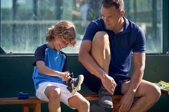 Focused man and cheerful boy in sportswear preparing to padel game and sitting outside against fence