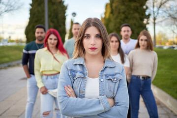 Self assured young female with crossed arms in denim jacket looking at camera while standing on street against friends on city park