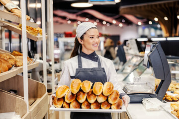 A happy saleswoman at bakery in supermarket holding tray with pastry.