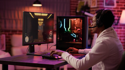 Handheld shot of african american man using pc gaming setup relaxing playing first person shooter in multiplayer. Gamer streaming while explaining gameplay in online action game talking to team.