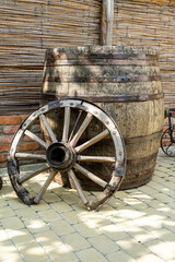 An old wooden barrel for wine, standing on a stone tile, against the background of a fence made of old bricks and reeds and an old, broken cartwheel leaning against it